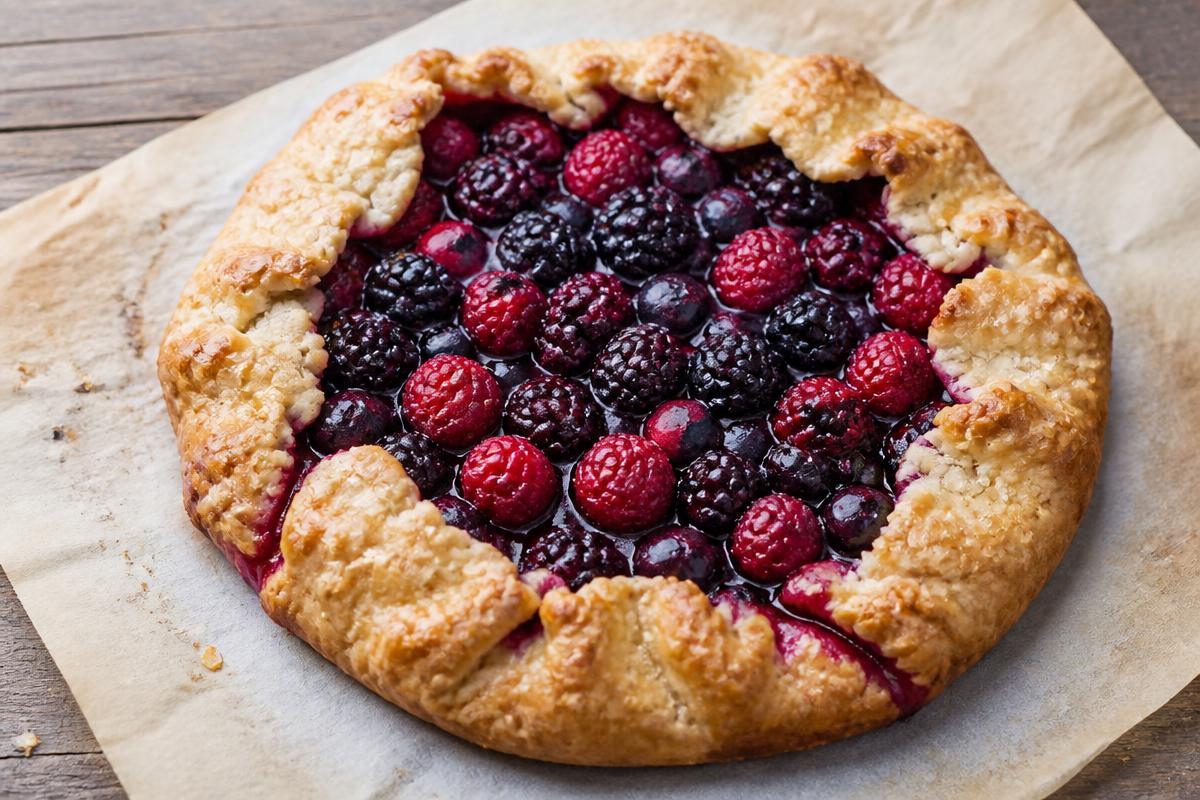 A rustic mixed berry galette on parchment with folded golden pastry around raspberries, blackberries and blueberries.